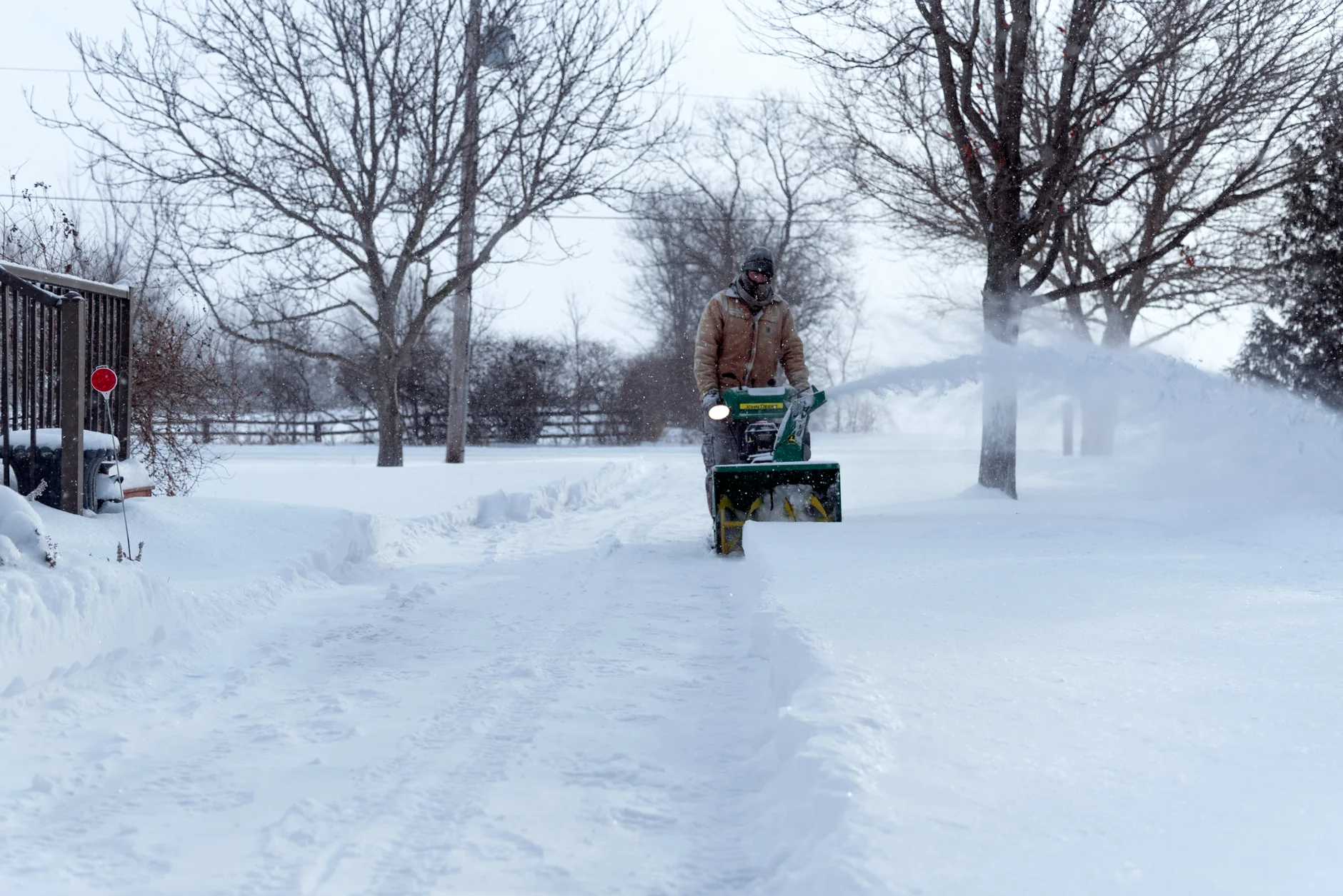 A person operates a snowblower on a snow-covered driveway in Essex, Ontario, during winter
